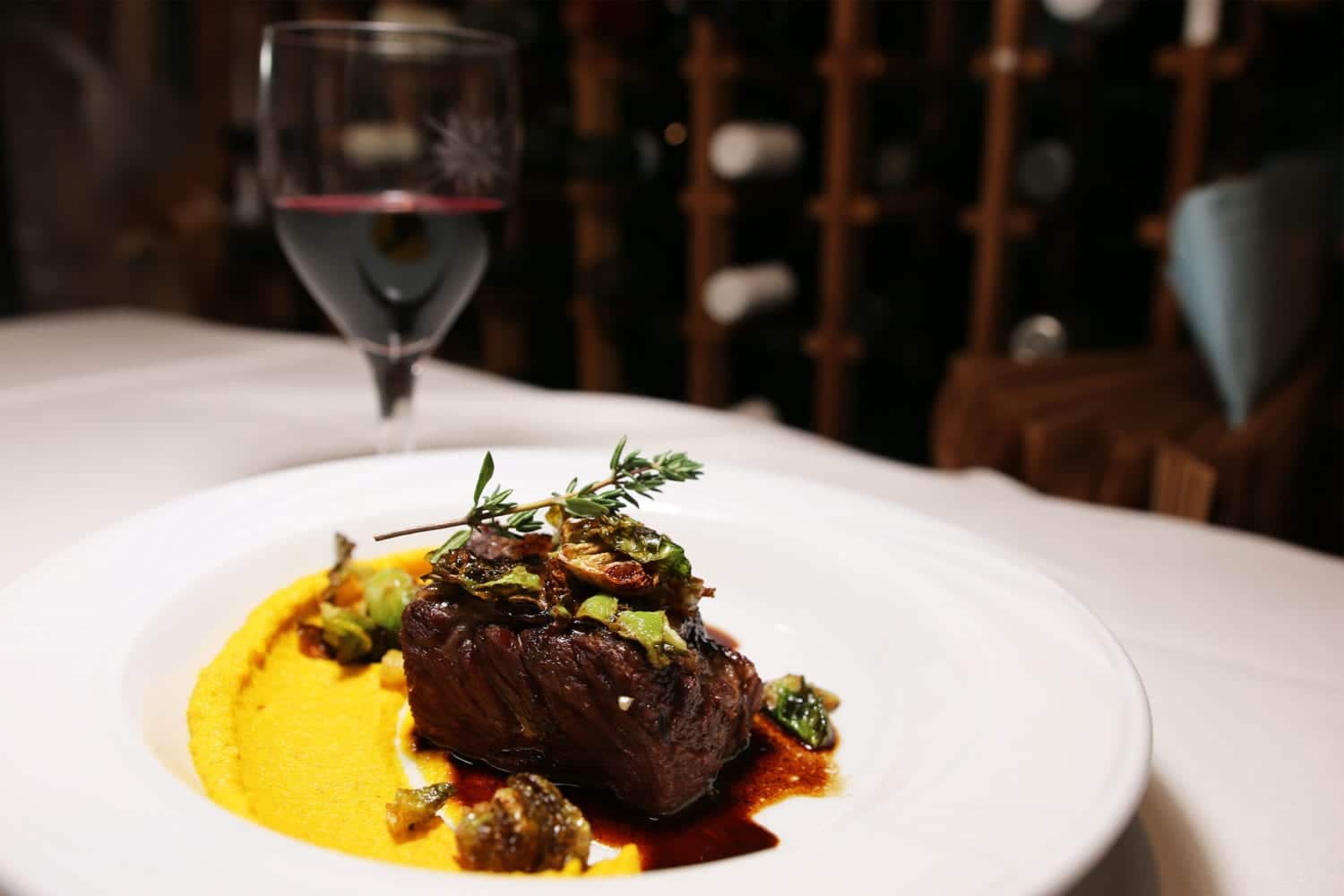 Beef Short Ribs topped with thyme on a white plate with wine glass in the background