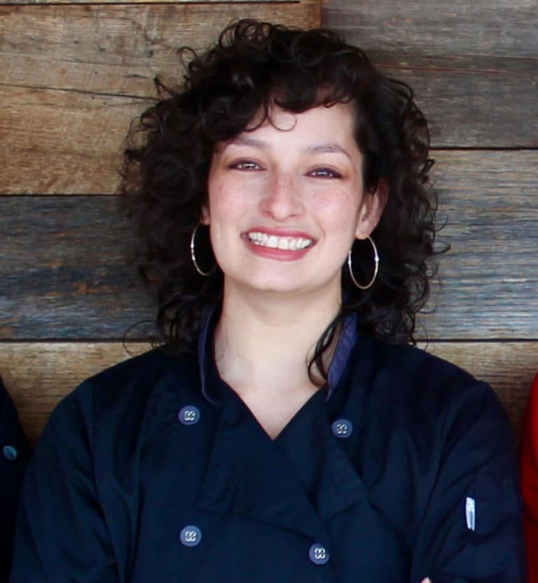 woman wearing denim chef coat leaning against a wood paneled wall